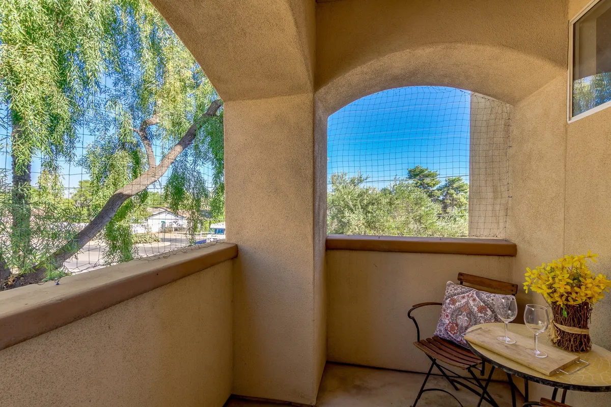 A private outdoor patio set with a small round table, two wine glasses, yellow flowers, a decorative pillow, and a view through the balcony overlooking palm trees. The soft lighting suggests either a morning or early evening ambiance, ideal for coffee or cocktails in a relaxed setting.