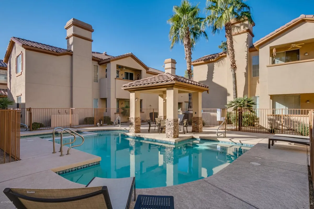 A resort-style community pool surrounded by palm trees and cabanas, with sun loungers and people swimming, sunbathing, and socializing. The pool area is framed by the backdrop of the community's buildings, emphasizing a casual and leisurely atmosphere.