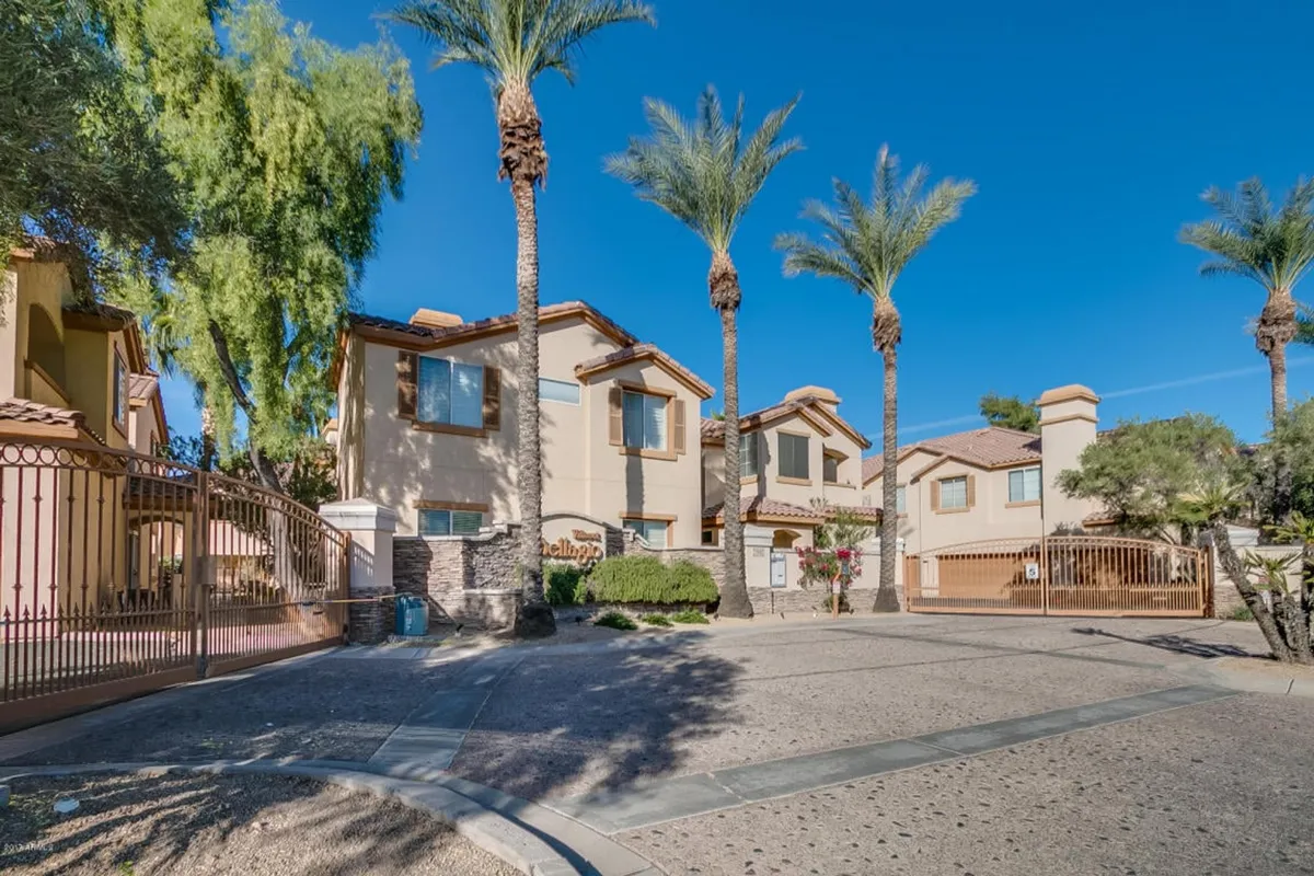 This exterior image features palm trees swaying above a welcoming entrance gate and landscaped garden. The building facade is partially visible, set against a tranquil neighborhood backdrop, inviting residents to step outside and explore the walkable community.