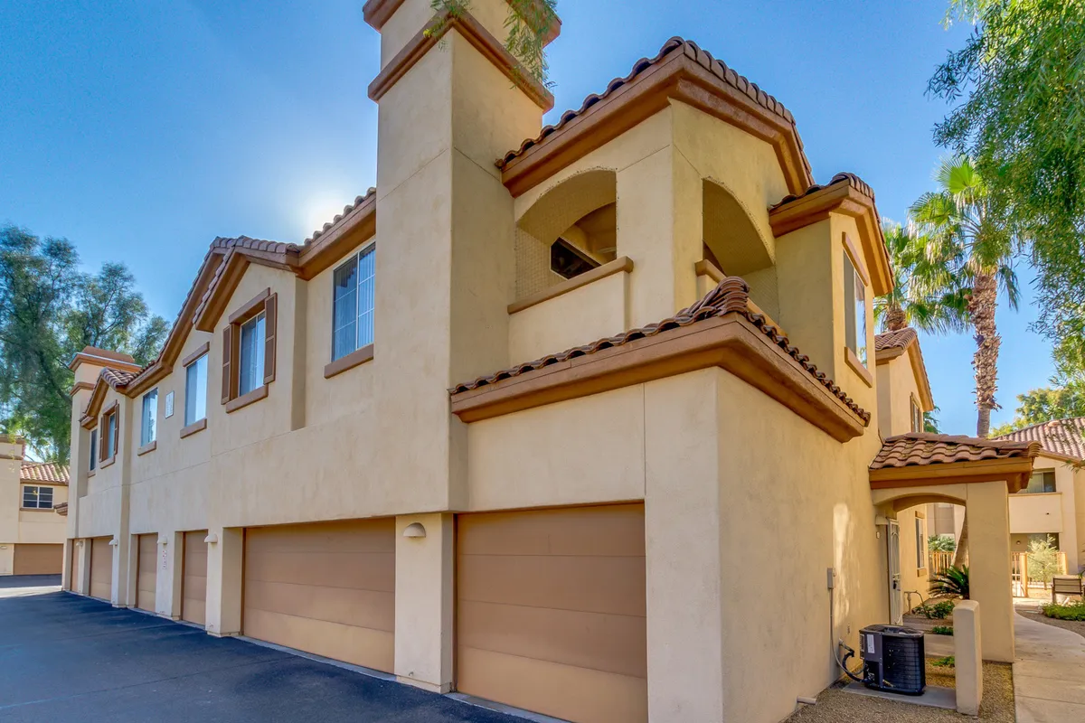 A warm, inviting exterior scene with textured stucco walls, classic decorative roof tiles, and sun-drenched palm trees. Spacious driveways and hints of a lively neighborhood suggest community activity and easy access to the vibrant surroundings.