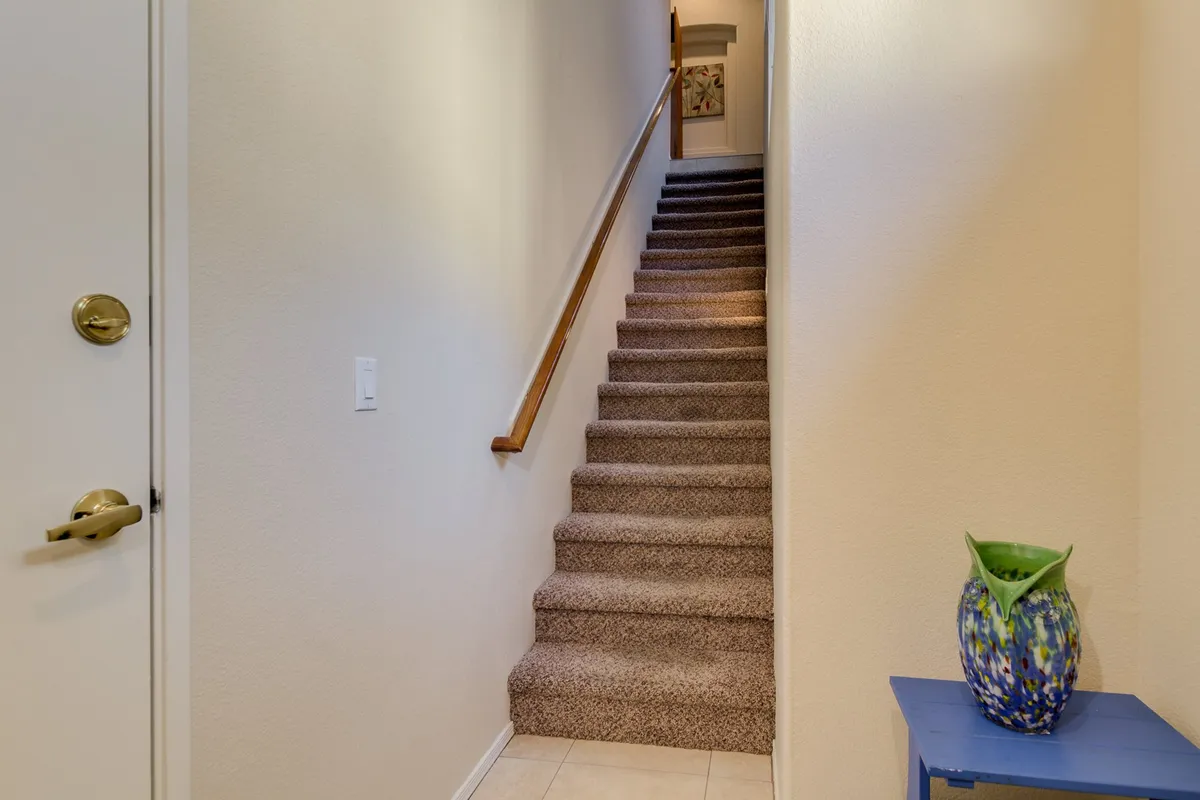An inviting interior shot showing a decorative staircase ascending from the foyer, with a blue table and vase adding character. The design highlights the transition from the entry level to the main living area, aligning directly with the narrative of ascending from the private garage/foyer to the home’s core spaces.